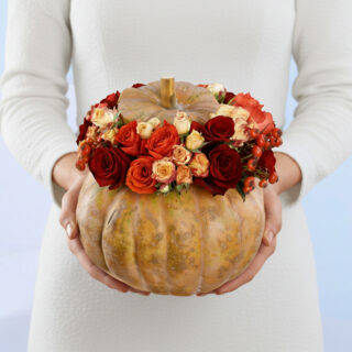 A person holding a large orange pumpkin filled with vibrant autumnal flowers. The pumpkin is topped with a smaller pumpkin lid, and the arrangement includes red, orange, and yellow roses, as well as small red berries. The background is a light blue.