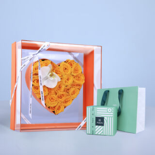 Heart-shaped arrangement of yellow roses and an orchid in a clear box with Patchi chocolate and shopping bag.