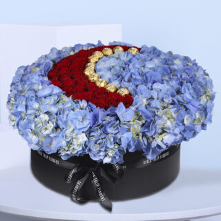 A round floral arrangement of blue hydrangeas, red roses, and gold chocolates, shaped like a crescent moon, in a black box with 'Black Tulip Flowers' branding