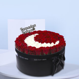 A floral arrangement of red and white roses shaped like a crescent moon in a black box, with a 'Ramadan Kareem' sign.