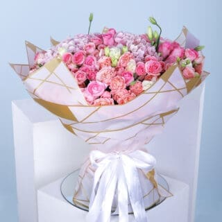 A bouquet of hydrangeas, baby roses, lisianthus, and pink roses wrapped in white with a gold border and a white ribbon.