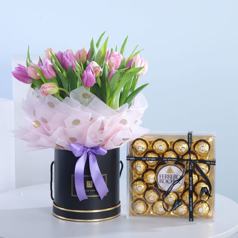 A bouquet of pink and purple tulips in a black hat box with a lilac ribbon, next to a box of Ferrero Rocher chocolates