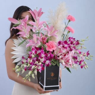 A floral arrangement with 6 pink lilies, 20 Dendrobium orchids, 5 pink carnations, and lush green leaves.