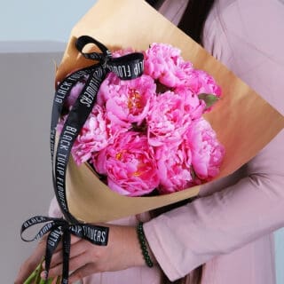 A person holding a bouquet of pink peonies wrapped in brown paper with a black ribbon.