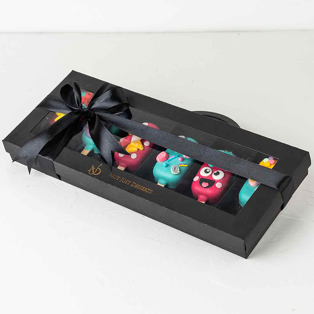 A black gift box with a clear window, tied with a black ribbon. Inside, there are several colorful cake pops on sticks, arranged in a row. The cake pops are decorated with various fun and whimsical designs. The box has the label "Not Just Desserts."
