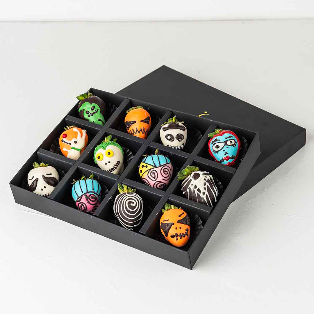 A black gift box filled with twelve chocolate-covered strawberries decorated with Nightmare Before Christmas characters. The characters include Jack Skellington, Sally, Zero, and other iconic figures.