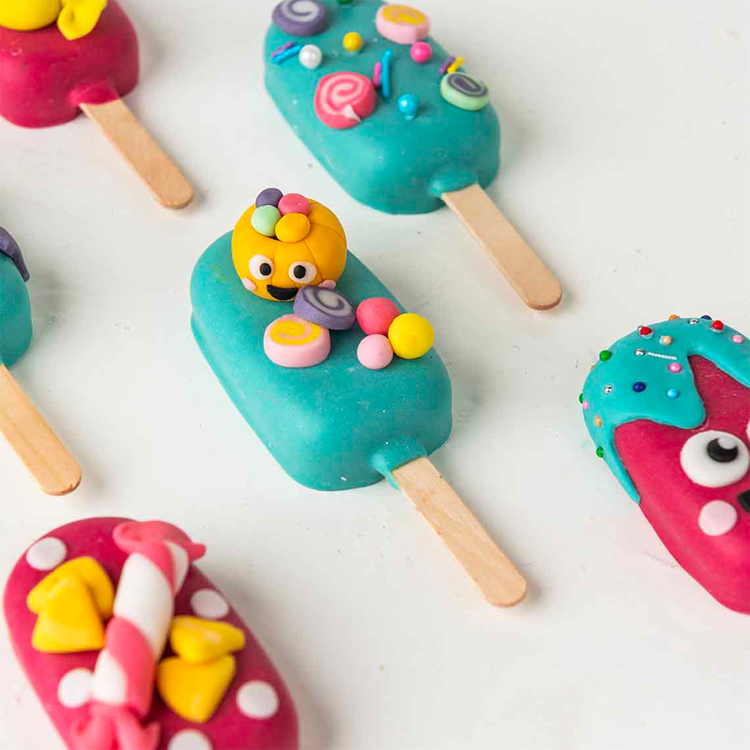 A close-up photo of several colorful cake pops on sticks. The cake pops are decorated with various fun and whimsical designs, including candy, sprinkles, and cartoon characters. One of the cake pops features a cute, smiling face made of fondant.
