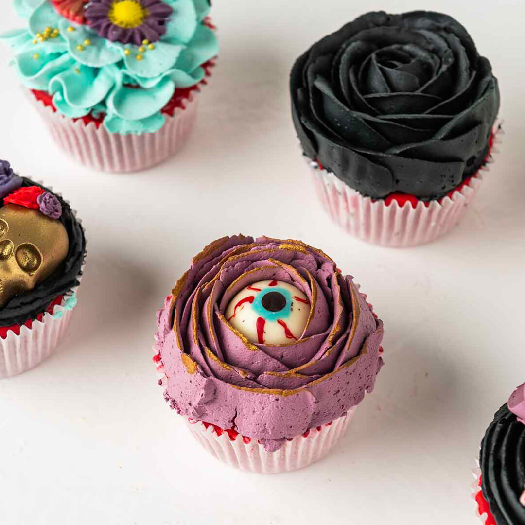 A close-up photo of a Halloween-themed cupcake. The cupcake is frosted with purple icing and decorated with a red and white eye in the center. The surrounding cupcakes are decorated with flowers, skulls, and other spooky designs.