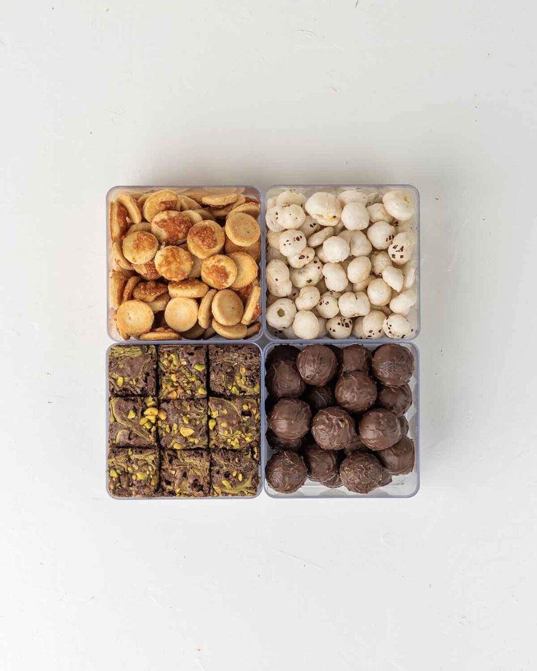 Four clear containers filled with different snacks. The top left container holds round, golden-brown snacks. The top right container holds white, round snacks. The bottom left container holds small, square brownies with nuts. The bottom right container holds round, dark chocolate truffles.