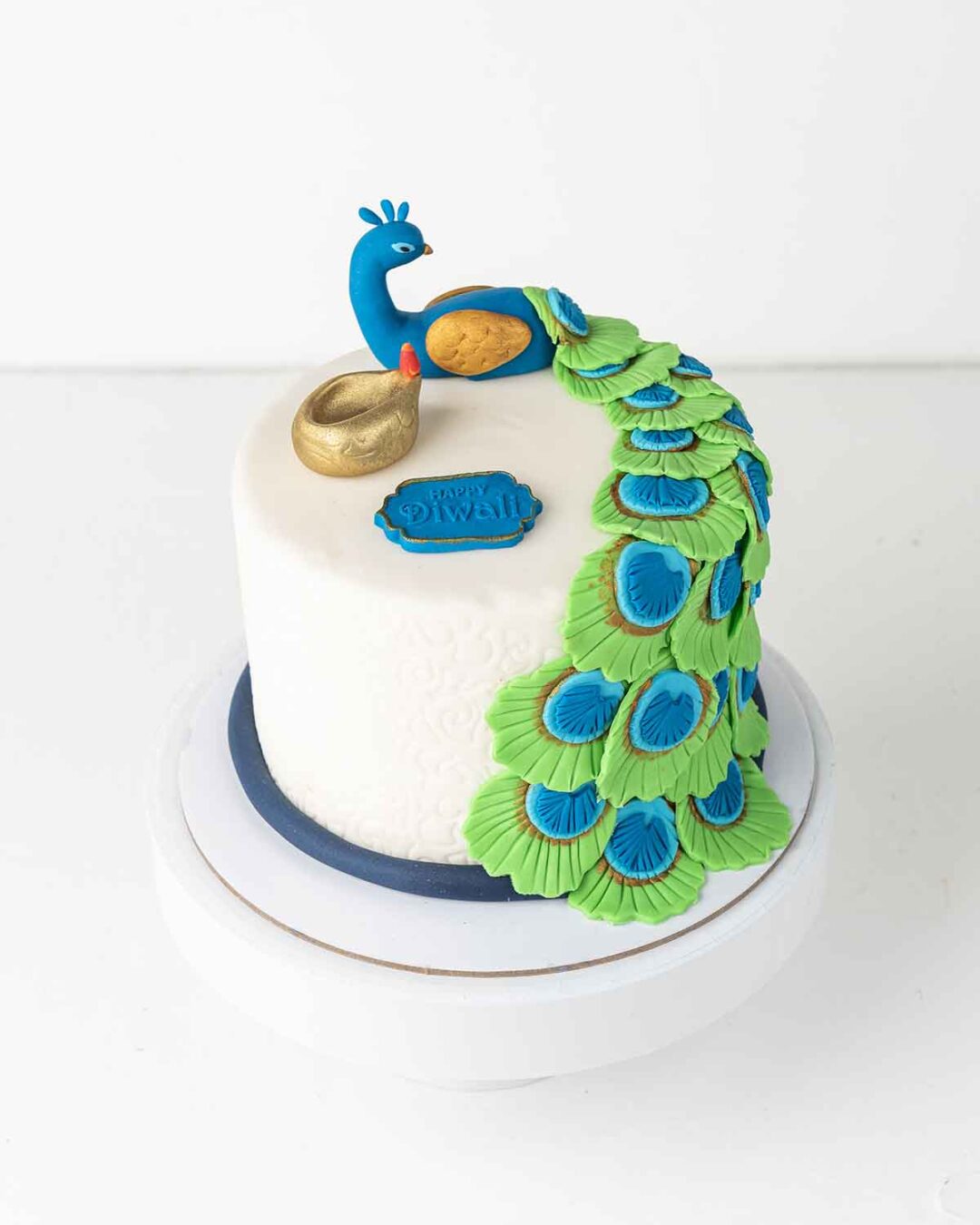 A white cake decorated with a blue and green peacock, a golden diya, and a sign that says Happy Diwali.