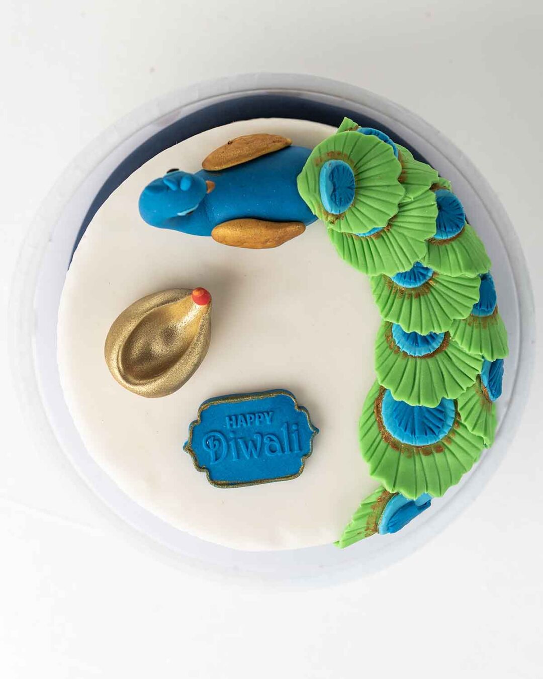 A white cake decorated with a blue and green peacock, a golden diya, and a sign that says "Happy Diwali.