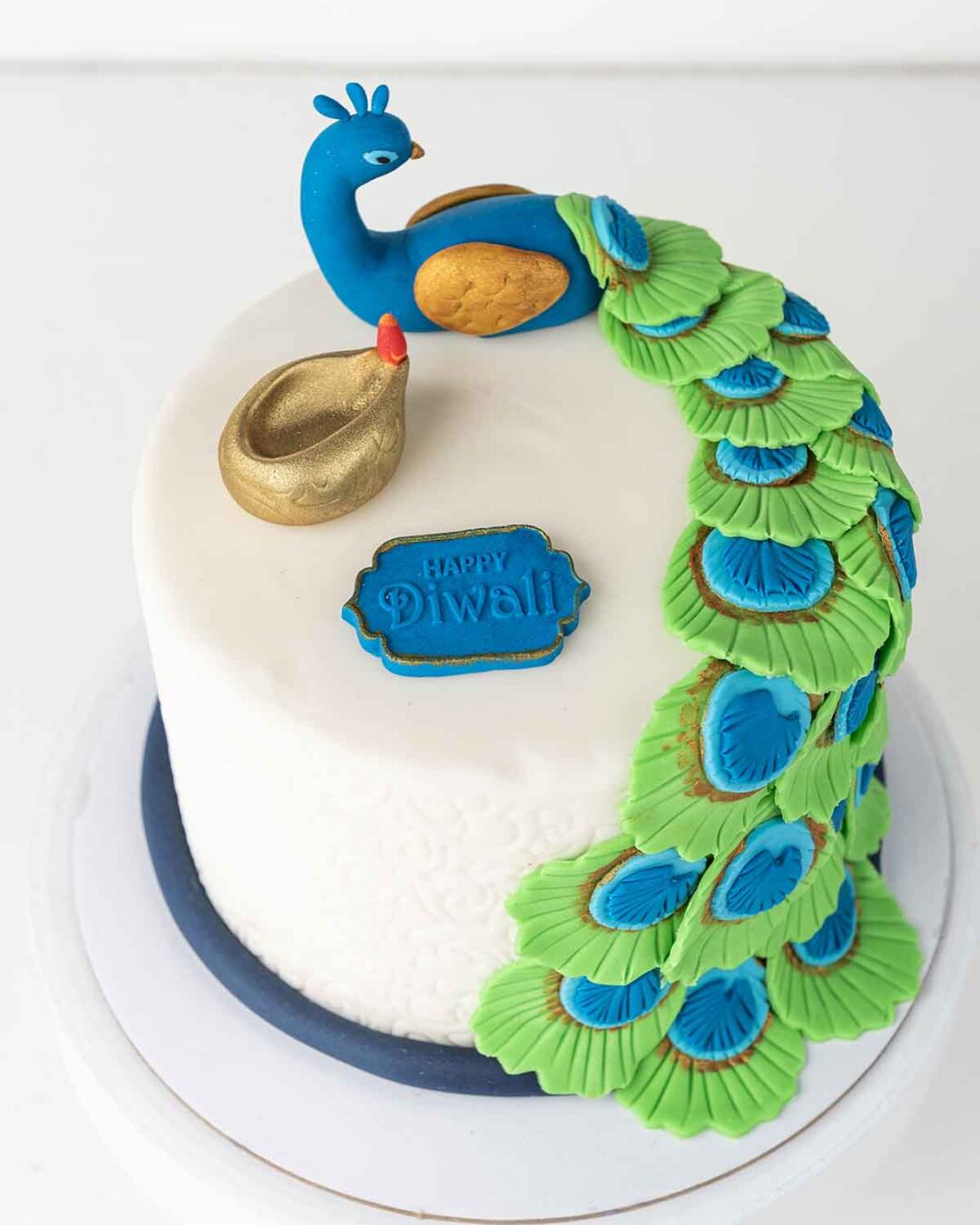 A white cake decorated with a blue and green peacock, a golden diya, and a sign that says "Happy Diwali.