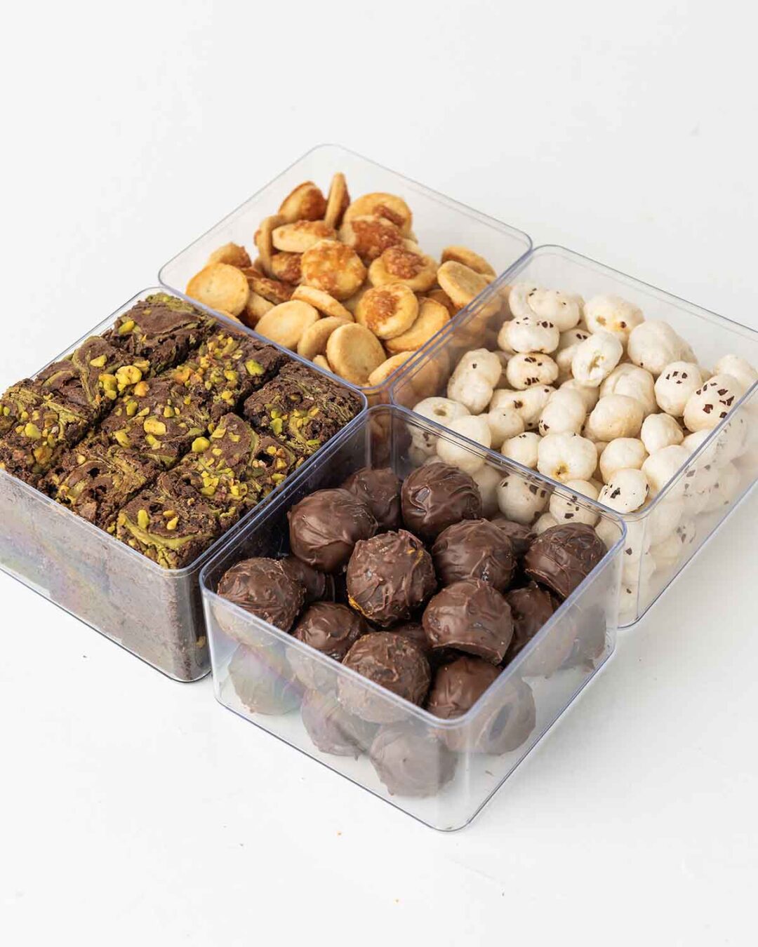 Four clear containers filled with different snacks. The top left container holds round, golden-brown snacks. The top right container holds white, round snacks. The bottom left container holds small, square brownies with nuts. The bottom right container holds round, dark chocolate truffles.