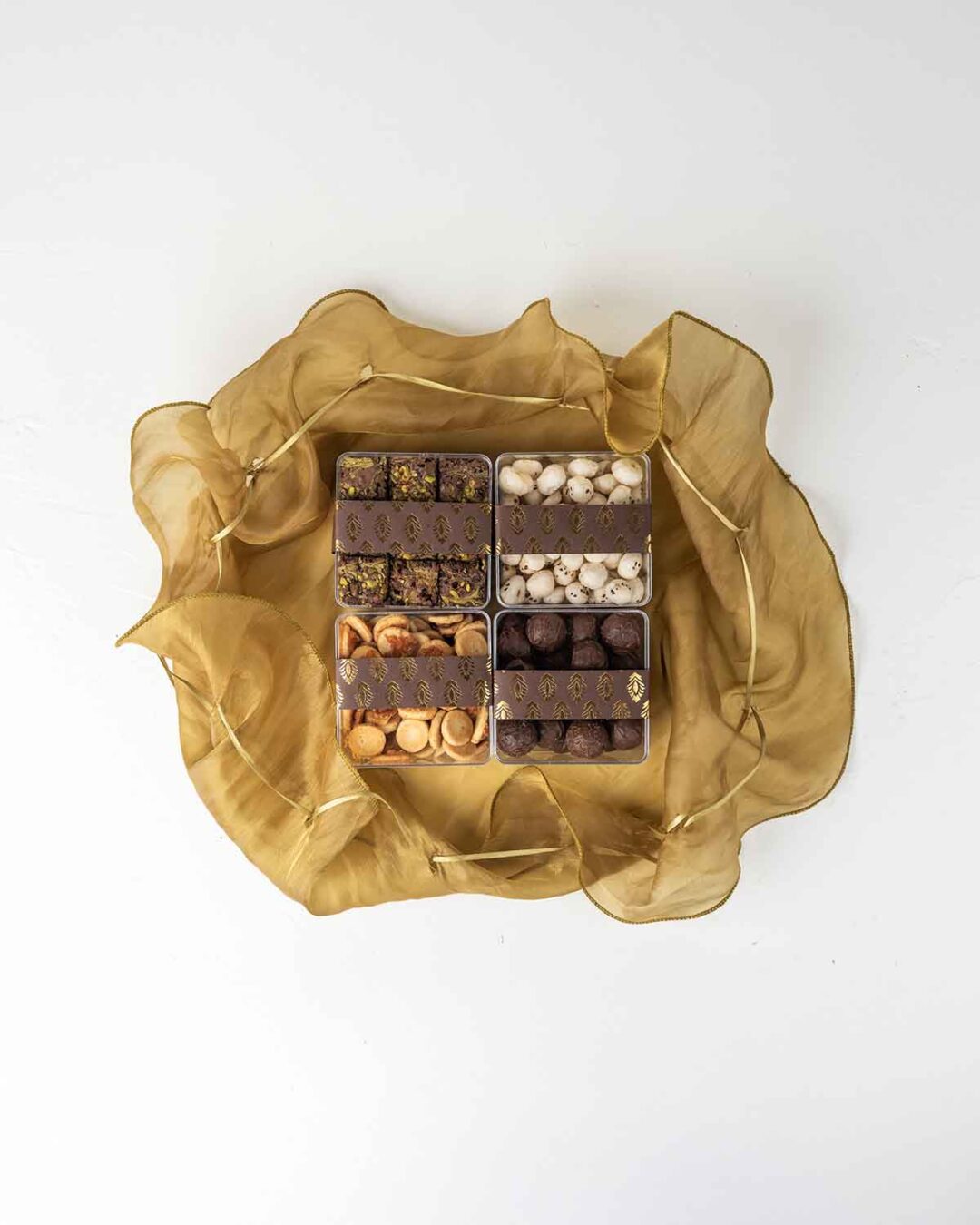 Four clear containers filled with different snacks arranged in a square basket lined with gold fabric. The containers hold various treats, including round, golden-brown snacks, white, round snacks, small, square brownies with nuts, and round, dark chocolate truffles.
