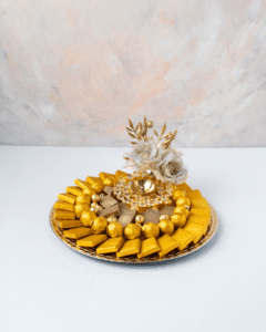 A circular silver platter filled with gold-wrapped chocolates. In the center of the platter is a small golden bowl with white flowers and a string of beads.