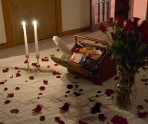 A romantic indoor picnic scene with a wicker basket filled with food, a vase of red roses, two lit candles, and scattered rose petals on a white sheet.
