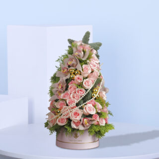 A festive tree arrangement with blush pink roses, orchids, golden ornaments, pearls, and greenery, set in an elegant pink base