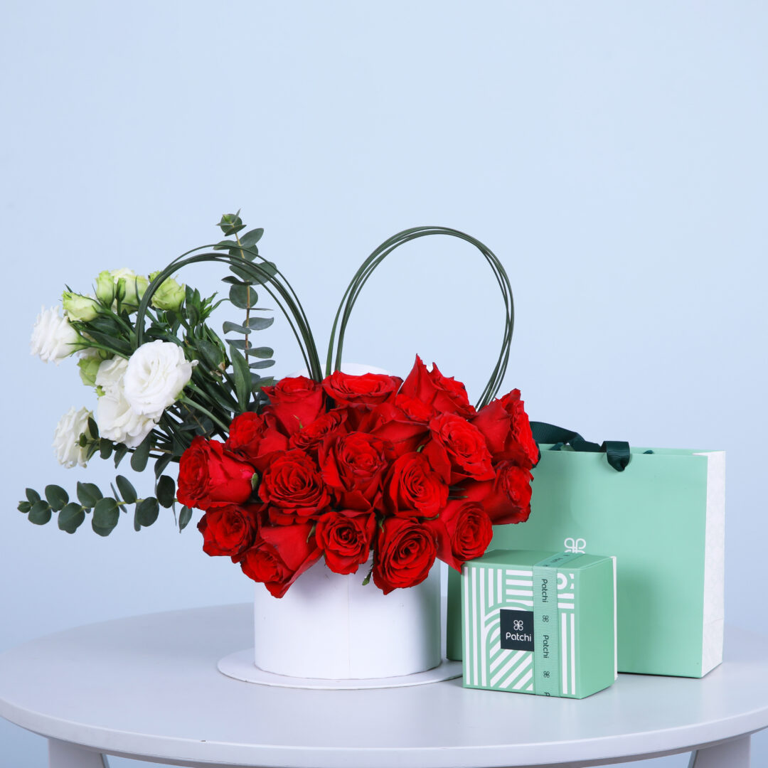 A white hat box filled with red, white and green roses accompanied with eucalyptus with a Patchi box.