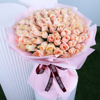 An elegant bouquet featuring fresh peach roses wrapped in blush pink paper and tied with a maroon ribbon.