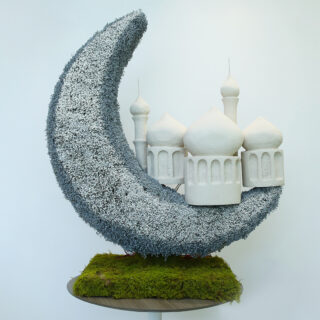 A grand crescent-shaped grey floral arrangement with a miniature white mosque, set on a moss-covered base on top of a sleek white stand.
