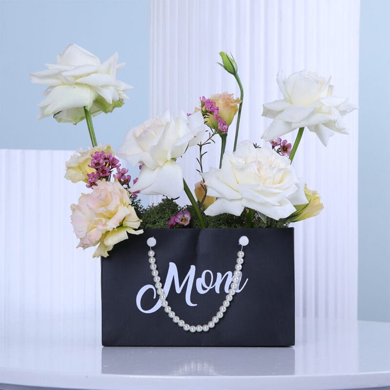 02 A gift set featuring white roses in a black Mom" planter, a green gift bag,and a box of Patchi chocolates