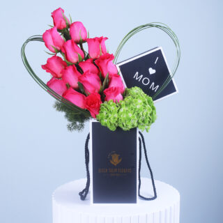 A heart-shaped floral arrangement with pink roses and green hydrangeas in a Black Tulip Flowers gift bag, featuring an "I Lov MOM" sign.