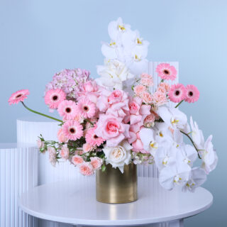 A floral arrangement with pink roses, pink gerberas, white orchids, and hydrangea in a gold vase.