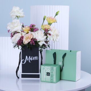 Pastel blooms arranged in a black 'Mom' gift box, paired with a green box of Patchi chocolates and a green gift bag.