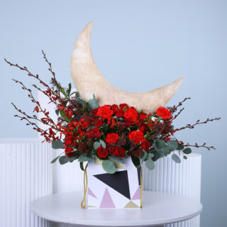 Red carnations with delicate branches in a geometric patterned box, with a textured crescent moon, displayed on a white table.