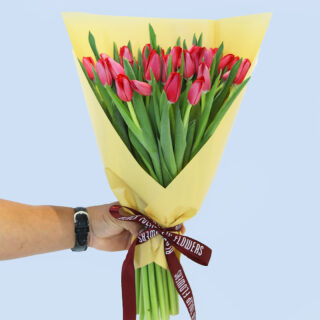 A hand holding a bouquet of red tulips wrapped in yellow paper and tied with a maroon ribbon.
