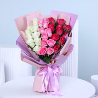 A bouquet of white, pink, and red roses clustered and wrapped in layers of light pink and lavender paper, tied with a wide, light purple satin ribbon.