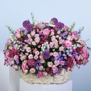 A pastel mixed flower basket arrangement featuring purple roses, pink baby roses, purple wax, delphinium, statice, and allium. The basket adds a rustic touch, making this arrangement an ideal gift for someone special.