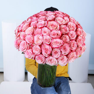 A large bouquet of 100 vibrant two-tone pink roses, with petals from a lighter pink at the center to a deeper, more saturated pink at the edges, is presented in a tall, clear glass cylindrical vase.