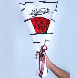 Bouquet of red roses wrapped in a white "Happy Anniversary" wrapper with a maroon ribbon, displayed on a white pedestal.