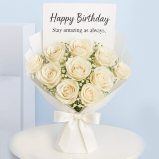 A bouquet of Cream roses with delicate baby's breath, wrapped in white paper and tied with a satin ribbon, with a "Happy Birthday, Stay amazing as always." card.