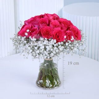 A vibrant bouquet of around 20 dark pink roses, surrounded by a cloud of white baby’s breath, is arranged in a clear glass vase with visible green stems tied with natural twine.