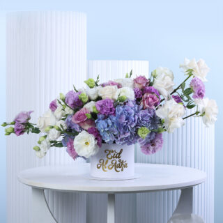 A luxurious floral arrangement featuring an assortment of purple and blue hydrangeas, white and light purple roses, and mixed blooms, presented in a white round box with "Eid Al Adha" written in gold script.