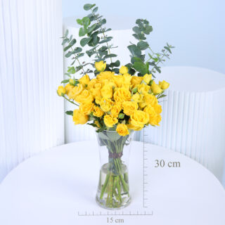 A bouquet of around 20 yellow spray rose stems, filled with bright blooms and lush green eucalyptus leaves, arranged in a clear glass vase with visible stems tied neatly with a brown ribbon.