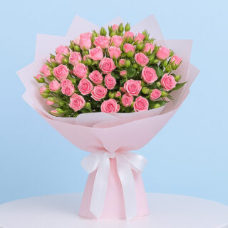 A lush bouquet of small, delicate pink spray roses and green buds wrapped in light pink with a slightly lighter pink inner layer and tied with a white ribbon