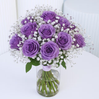 A luxurious bunch of purple roses with baby’s breath in a clear glass vase.
