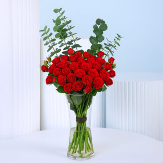 20 vibrant red spray roses in a clear glass vase adorned with green eucalyptus leaves.