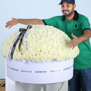 A man in a green polo shirt carefully places a single white rose into a very large, round floral arrangement made entirely of white roses.