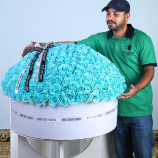 A man in a green polo shirt gestures towards a very large, round floral arrangement made entirely of vibrant aqua blue roses in a white box.