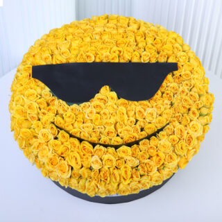 A large, round floral arrangement filled with yellow roses with black "sunglasses" emoji in a black box.