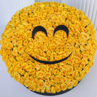A large floral arrangement shaped like a yellow smiling emoji, made entirely of yellow roses. It has two black curved shapes for eyes and a black curved line for a smile presented in a black round box.