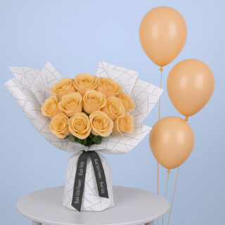 A bouquet of peach roses arranged in a white wrap with a black geometric design. It is tied with a black ribbon accompanied with peach-colored balloons.