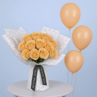 A bouquet of peach roses arranged in a white wrap with a black geometric design. It is tied with a black ribbon accompanied with peach-colored balloons.