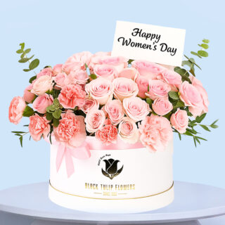 A round White box filled with pink roses and carnations with a small message card with the text "Happy Women's Day".