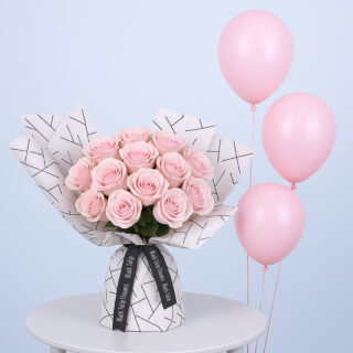 A bouquet of light pink roses wrapped in white paper with a black geometric pattern along with pink balloons.