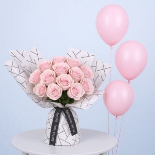 A bouquet of light pink roses wrapped in white paper with a black geometric pattern along with pink balloons.